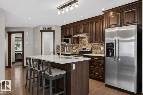 Kitchen featuring dark wood-finish cabinetry, a central island with a white countertop, a stainless steel side-by-side refrigerator, a stainless steel range with overhead microwave, and stone tile backsplash - 8506 80 Avenue, Edmonton, AB 
