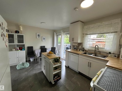Kitchen featuring dark gray tile flooring, white cabinetry, light wood-finish countertops, a double basin stainless steel sink with a high-arch faucet, and a sliding glass door - 10924 30 Street, Edmonton, AB 