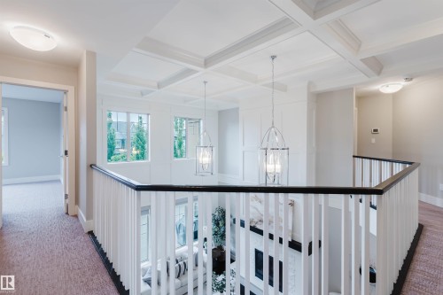 Upper-level hallway overlooking a two-story space, featuring white coffered ceilings, modern pendant lighting, and a white baluster railing with a dark wood-finish handrail - 3435 Keswick Boulevard, Edmonton, AB 