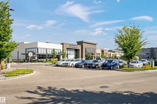 Modern commercial complex featuring a light-toned stucco facade, expansive storefront windows, a contemporary dark grey brick building, an asphalt parking lot, and mature trees with landscaped islands - 3435 Keswick Boulevard, Edmonton, AB 