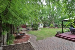 Expansive backyard featuring a brick paver patio, a raised wood deck with a pergola, a dedicated fire pit area, a storage shed, and mature trees providing natural shading - 