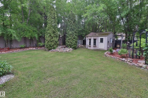 Expansive backyard featuring a manicured lawn, a detached storage shed with paneled siding, a defined garden bed with rock edging and mulch, mature trees along the perimeter, and a dark-finished arched trellis - 42 Alpaugh Crst, Leduc, AB 
