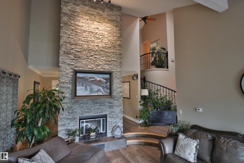 Vaulted living room featuring a floor-to-ceiling stacked stone fireplace with a raised hearth, wood-finish flooring, and a wrought iron staircase with an upper-level balcony - 42 Alpaugh Crst, Leduc, AB 