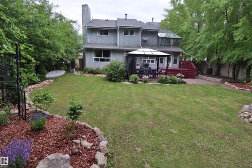 Expansive backyard featuring a manicured lawn, a red-stained wooden deck, a paved patio, a sunroom with an angled glass roof, and mature perimeter trees - 42 Alpaugh Crst, Leduc, AB 