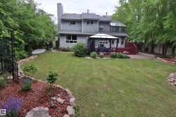 Expansive backyard featuring a manicured lawn, a red-stained wooden deck, a paved patio, a sunroom with an angled glass roof, and mature perimeter trees - 