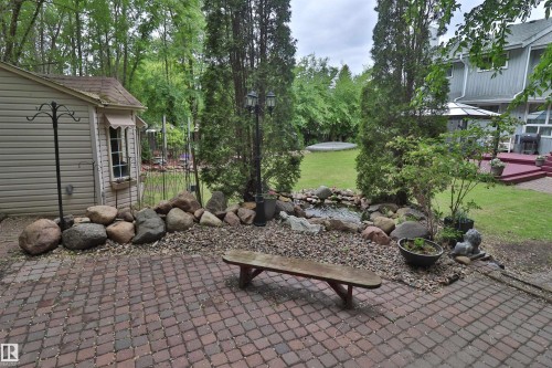 Paver patio extending to a landscaped rock garden with a water feature, bordered by mature trees and a manicured lawn - 42 Alpaugh Crst, Leduc, AB 