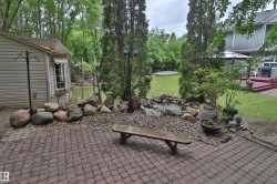 Paver patio extending to a landscaped rock garden with a water feature, bordered by mature trees and a manicured lawn - 