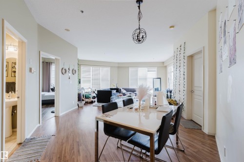 Open-concept living area with wood-finish flooring, a contemporary chandelier, and large windows with privacy blinds - 323 4831 104A Street, Edmonton, AB 