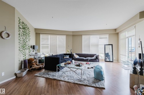 Spacious living area featuring wood-finish flooring, light-toned walls, and multiple large windows with blinds - 323 4831 104A Street, Edmonton, AB 