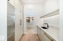 Contemporary galley-style kitchen with light wood-finish flooring, integrated stainless steel refrigerator, white cabinetry, and a black sink with a matte black faucet - 