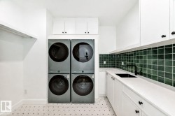 Laundry area featuring stacked washer and dryer units, white cabinetry, a black sink, and a dark green tile backsplash - 