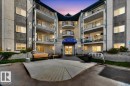 Multi-story building featuring stacked balconies with metal railings, a central entrance with a blue awning, and a concrete approach with a wooden bench - 112 6710 158 Avenue, Edmonton, AB 