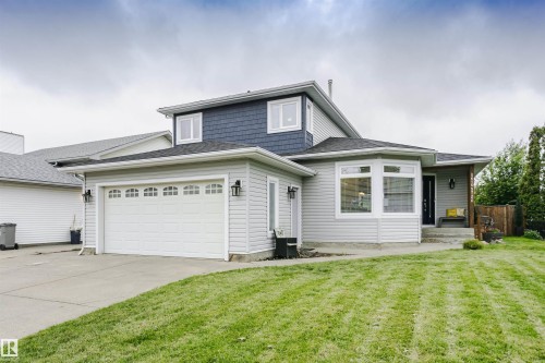 Two-story residence featuring a two-tone exterior with light-colored siding and dark blue shingles, an attached two-car garage with a concrete driveway, a front entry with a bay window, and a sloped front lawn - 5610 57 Avenue, Beaumont, AB 