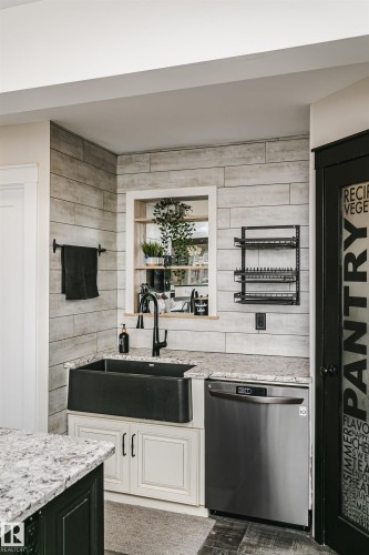 Kitchen featuring a black farmhouse sink, matte black faucet, light granite countertops, stainless steel dishwasher, and shiplap-style wall paneling - 5610 57 Avenue, Beaumont, AB 