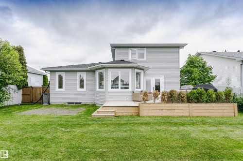 Expansive backyard featuring a manicured lawn, a multi-level wood deck, a wood planter box with greenery, light gray siding, and multiple white-framed windows - 5610 57 Avenue, Beaumont, AB 