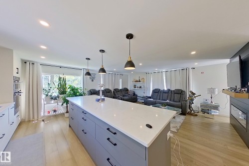 Open-concept common area featuring a spacious kitchen island with a light-colored countertop and dark-finish hardware, recessed lighting, wood-finish flooring, a wall-mounted double oven, and pendant lighting fixtures - 3054 28 Avenue, Edmonton, AB 