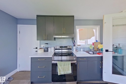 Kitchen featuring gray base cabinetry and green upper cabinetry with gold-toned hardware, white solid surface countertops, and stainless steel appliances - 3054 28 Avenue, Edmonton, AB 