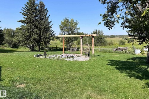 Expansive backyard featuring a circular fire pit with stone surround, a wooden swing structure, and mature evergreen trees - 3054 28 Avenue, Edmonton, AB 