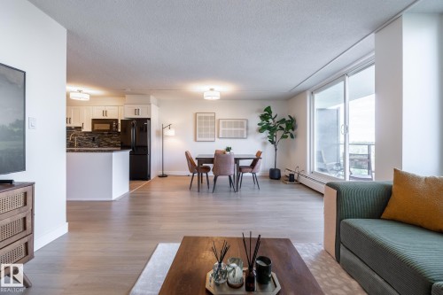 Expansive living area with wood-finish flooring, featuring a kitchen with white cabinetry and a dark tile backsplash, a dedicated dining area, and a large sliding glass door leading to a private outdoor space - 807 10011 116 Street, Edmonton, AB 