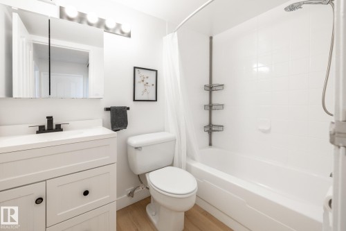 Bathroom featuring a white vanity with black hardware, a matte black faucet, a mirrored medicine cabinet, a white bathtub with tile surround, and wood-finish flooring - 807 10011 116 Street, Edmonton, AB 