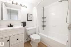 Bathroom featuring a white vanity with black hardware, a matte black faucet, a mirrored medicine cabinet, a white bathtub with tile surround, and wood-finish flooring - 