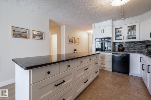 Kitchen featuring a large island with dark stone countertop, white cabinetry with black hardware, integrated wall oven, black dishwasher, and tiled backsplash - 807 10011 116 Street, Edmonton, AB 