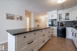 Kitchen featuring a large island with dark stone countertop, white cabinetry with black hardware, integrated wall oven, black dishwasher, and tiled backsplash - 