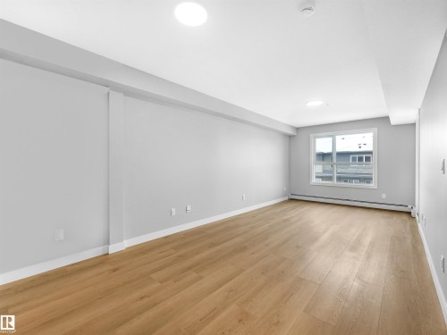 Spacious room featuring wood-finish flooring, light grey painted walls, white baseboards, and a large window with white framing - 318 1506 Tamarack Boulevard, Edmonton, AB 