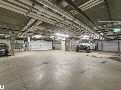 Underground parking garage featuring concrete flooring, exposed ceiling utilities, concrete support columns, dedicated storage units with mesh doors, and a roll-up vehicle access door - 318 1506 Tamarack Boulevard, Edmonton, AB 