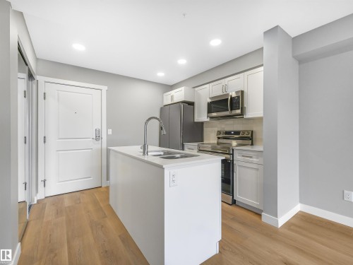 Kitchen featuring a central island with a sink, stainless steel appliances, white cabinetry, light countertops, and wood-finish flooring - 318 1506 Tamarack Boulevard, Edmonton, AB 