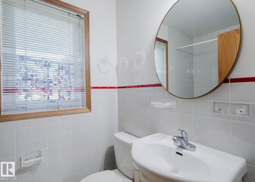 Bathroom featuring white ceramic wall tiles with a red accent band, a white pedestal sink with a chrome faucet, and a round wall-mounted mirror - 10948 75 Avenue, Edmonton, AB 