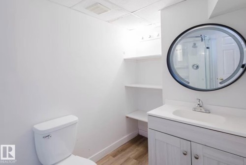 Bathroom featuring a round mirror, single vanity with integrated sink, wood-finish flooring, built-in shelving, and a toilet - 10948 75 Avenue, Edmonton, AB 