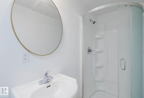 Bathroom featuring a pedestal sink with chrome faucet, a circular mirror with a gold-tone frame, and a curved stand-up shower with integrated shelving - 10948 75 Avenue, Edmonton, AB 