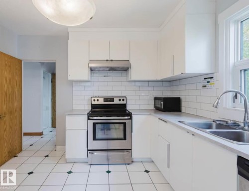 Bright kitchen featuring white cabinetry, subway tile backsplash, stainless steel range with overhead hood, double basin sink, and tile flooring with accent squares - 10948 75 Avenue, Edmonton, AB 