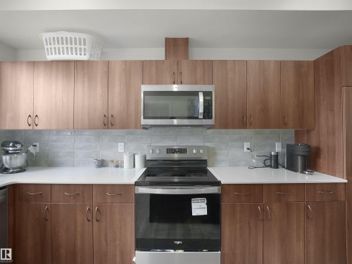 Modern kitchen featuring wood-finish cabinetry, stainless steel appliances, a white countertop, and a light gray tile backsplash - 5022 Kinney Link Sw, Edmonton, AB 