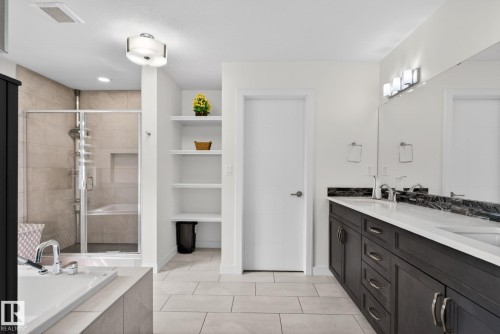 Spacious bathroom featuring a built-in soaking tub, glass-enclosed shower with tiled walls, dual dark wood-finish vanities with white countertops and undermount sinks, and integrated shelving - 704 39 Street, Edmonton, AB 
