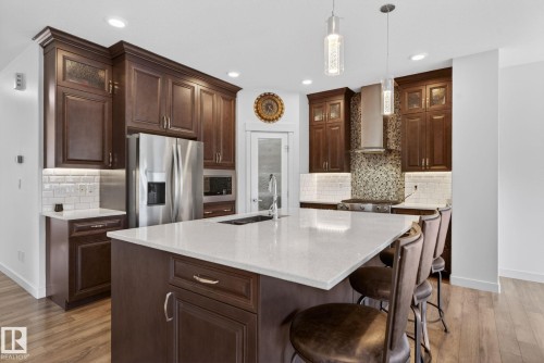 Contemporary kitchen featuring a large central island with seating, dark wood cabinetry, stainless steel appliances, and wood-finish flooring - 704 39 Street, Edmonton, AB 