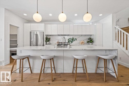 Contemporary kitchen featuring a substantial island with stone countertop and integrated sink, stainless steel appliances, sleek white cabinetry, a stone slab backsplash, and light wood-finish flooring - 10641 126 Street, Edmonton, AB 