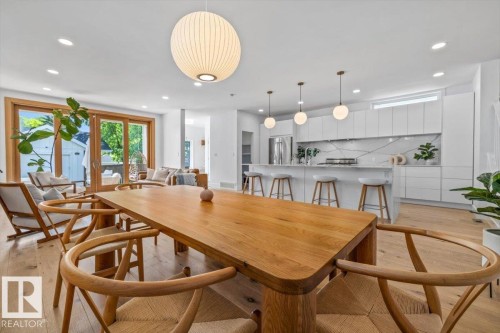 Open-concept living area featuring light wood-finish flooring, a contemporary dining table, a modern kitchen with white cabinetry, a large kitchen island with seating, and expansive glass doors opening to the exterior - 10641 126 Street, Edmonton, AB 