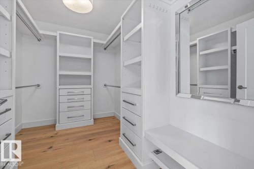 Custom closet system featuring wood-finish flooring, white built-in shelving, integrated drawers with horizontal pulls, multiple hanging rods, and a mirrored vanity area with a built-in bench - 10641 126 Street, Edmonton, AB 