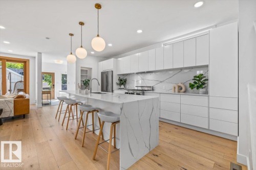 Sleek kitchen featuring a waterfall island with stone countertop, white flat-panel cabinetry, stainless steel appliances, and wood-finish flooring - 10641 126 Street, Edmonton, AB 
