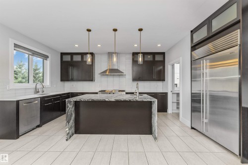 Modern kitchen featuring a waterfall island with stone countertop, dark wood-finish cabinetry, stainless steel appliances, white subway tile backsplash, and light-toned tile flooring - 3310 Watson Bay, Edmonton, AB 