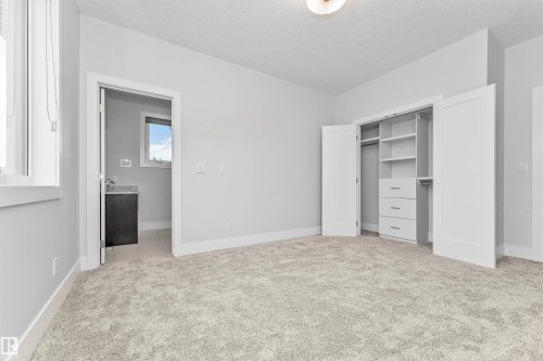 Spacious room featuring light gray walls, plush carpeting, a built-in closet system with shelving and drawers, a window with white trim, and a doorway revealing a vanity with a dark countertop and a small window - 3310 Watson Bay, Edmonton, AB 