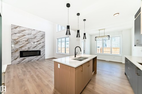 Open-concept living space featuring a central kitchen island with a double sink, a striking fireplace with a stone-finish facade, high ceilings, large windows, and wood-finish flooring throughout - 341 Roberts Wynd, Leduc, AB 