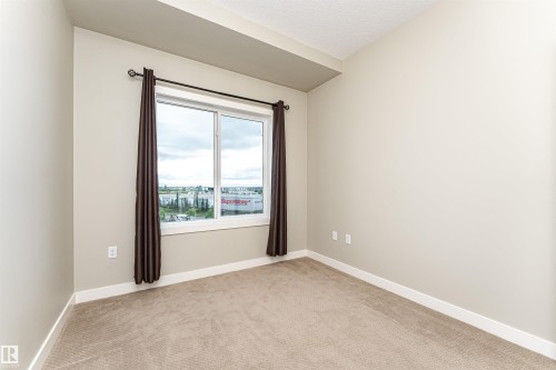 Window with a dark curtain rod and drapes, light beige walls, white baseboards, and tan carpet flooring - 505 1230 Windermere Way, Edmonton, AB 