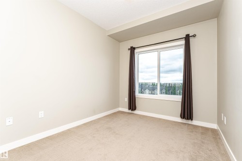 Neutral-toned room featuring a large window with white framing, beige carpeting, and white baseboards - 505 1230 Windermere Way, Edmonton, AB 