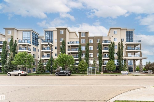 Multi-story building featuring a stucco and stone facade, multiple balconies with glass railings, a prominent covered entrance, and extensive landscaping with mature trees - 505 1230 Windermere Way, Edmonton, AB 