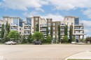 Multi-story building featuring a stucco and stone facade, multiple balconies with glass railings, a prominent covered entrance, and extensive landscaping with mature trees - 505 1230 Windermere Way, Edmonton, AB 