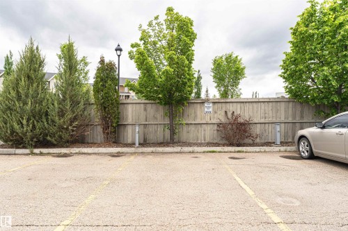 Paved parking area featuring painted yellow parking lines, a wooden privacy fence, mature trees, and a street light - 505 1230 Windermere Way, Edmonton, AB 