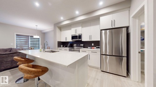 Modern kitchen featuring an island with a built-in sink, white cabinetry, integrated stainless steel appliances, a dark tile backsplash, and recessed lighting - 607 31 Avenue, Edmonton, AB 